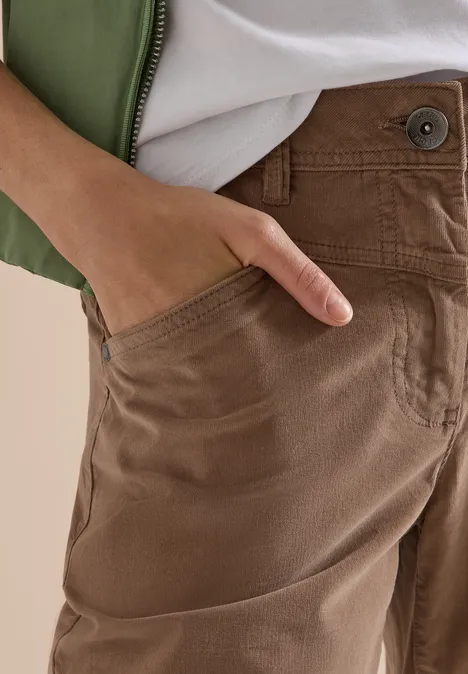 Gros plan: main dans la poche d'un pantalon marron, avec un manteau vert et un t-shirt blanc.
