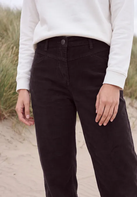 Eine Person steht im Sand, trägt weißen Pullover und schwarze Hose, im Hintergrund Dünen.