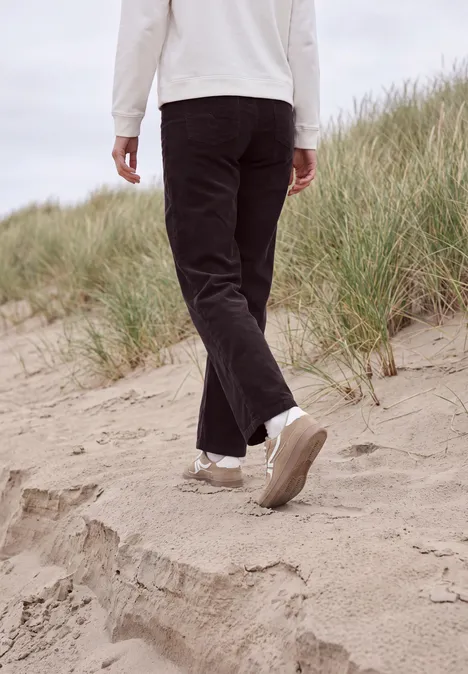 Eine Person in schwarzen Hosen und weißen Sneakern geht über sandige Dünen am Strand.