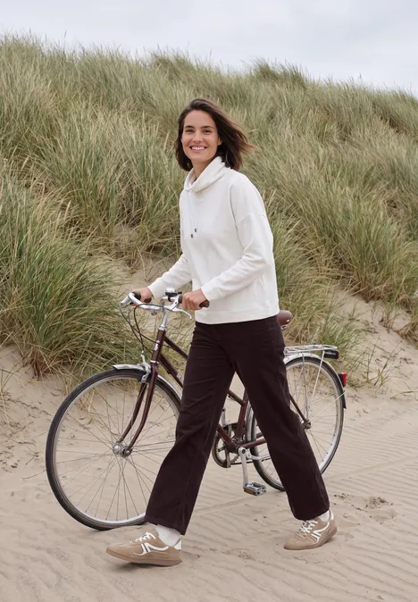 Eine Frau in weißem Pullover schiebt fröhlich ihr Fahrrad an grasigen Dünen entlang.