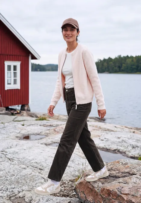 Frau in Jacke und Hose spaziert auf Felsen am See, rotes Haus im Hintergrund