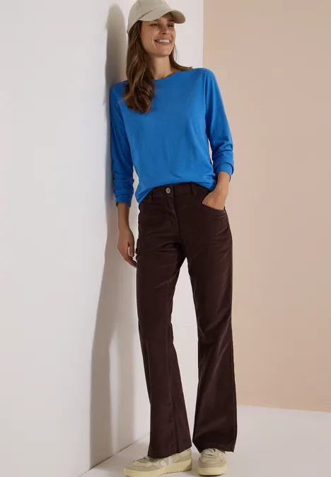 High Waist Bootcut Hose im Slim Fit truffle brown Eine Frau lehnt an einer Wand, trägt ein blaues Shirt, braune Hose und beige Baseballkappe, sie lächelt.