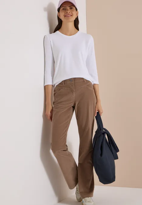 Une femme appuyée contre un mur porte un haut blanc, un pantalon marron, une casquette et un sac bleu foncé.