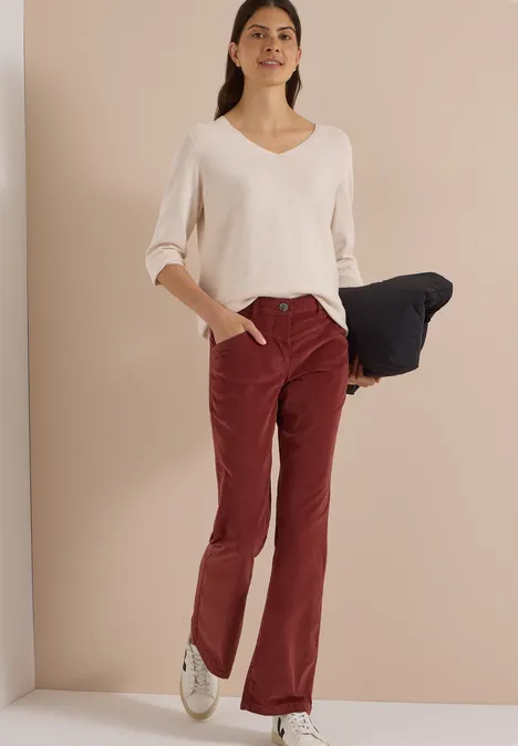 Femme en pull blanc à col en V et pantalon rouge, tenant un vêtement sombre, fond beige.