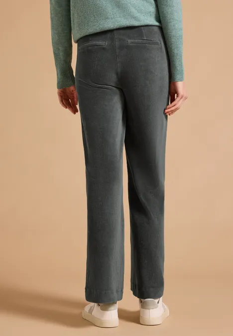 High Waist Straight Leg Hose w kroju Loose Fit easy khaki Widok z tyłu osoby w szarych sztruksowych spodniach, zielonym swetrze i białych sneakersach na beżowym tle.