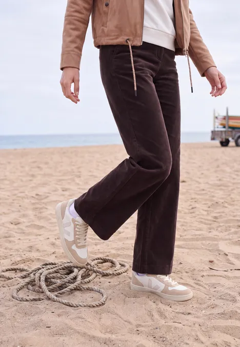 Frau in brauner Cord-Hose und weißen Sneakern am Strand stehend neben einem Seil