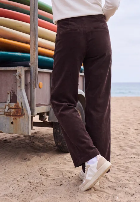 Person am Strand trägt braune Cordhose und weiße Sneaker, steht neben Van mit Surfboards.