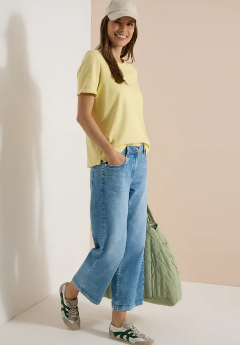 Frau in gelbem T-Shirt, weitem Jeans, Sandalen und grünem Stoffbeutel.