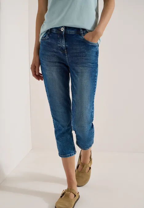 Eine Person trägt blaue, hochgekrempelte Jeans und beige Loafers vor weißem Hintergrund.