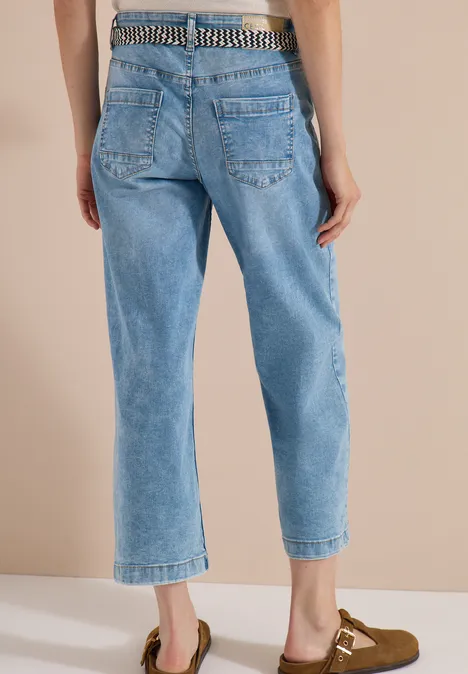 Vue arrière d’une personne en jean bleu clair court, ceinture à clous et mules marron.