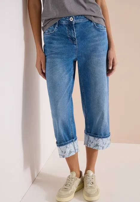 7/8 Wide Leg Jeans mit Turn-Up-Detail mid blue wash Person trägt dunkelblaue, knöchellange Jeans mit Umschlag und beige Sneaker vor heller Wand.