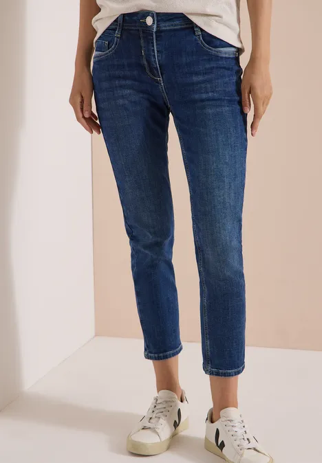 Mid Waist Slim Leg Jeans im Casual Fit mid blue used wash Eine Person trägt dunkelblaue, knöchellange Jeans und weiße Sneaker vor einem hellen Hintergrund.