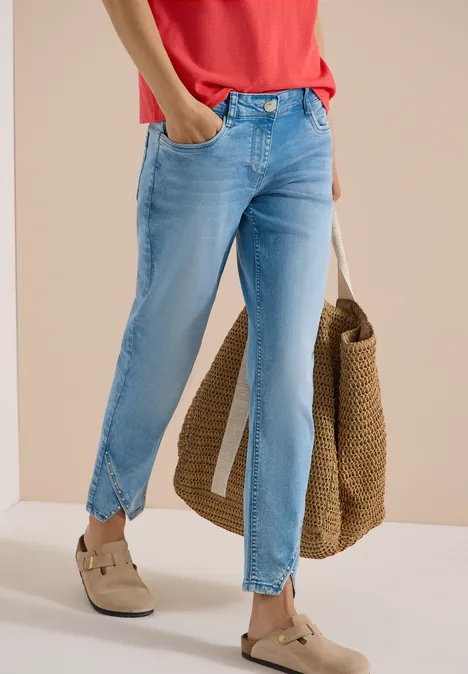Frau trägt hellblaue Jeans mit Knöchelschlitz, rotes Shirt, Basttasche und helle Slipper.