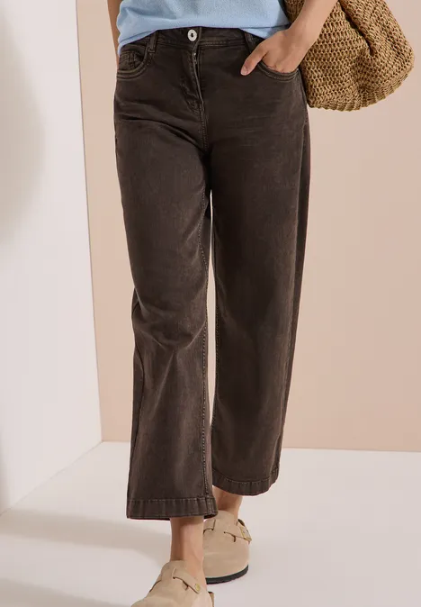 7/8 High Waist Jeans im Loose Fit brown cacao Frau trägt weite dunkelbraune Cordhose mit weißen Slippern und Basttasche.