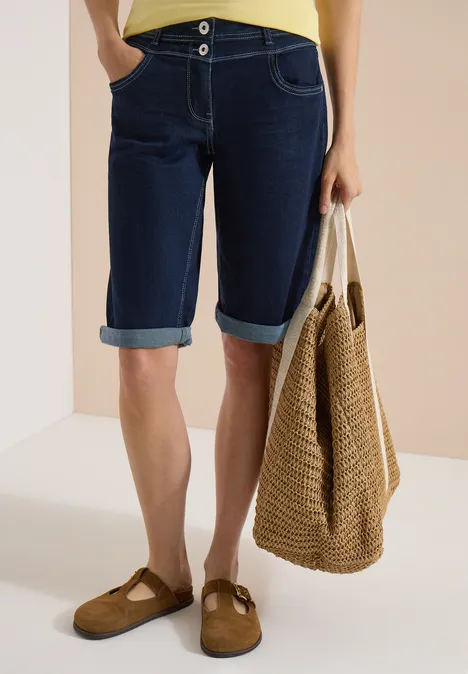 Beeld van benen in knielange spijkershorts, met een rieten tas en bruine muiltjes.