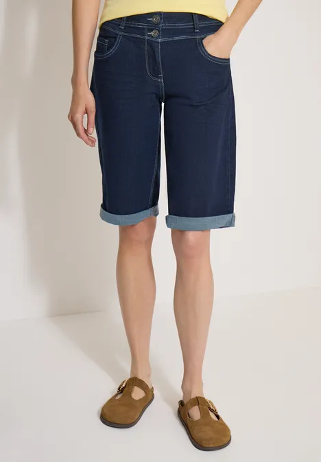 Persoon in donkere, knielange jeansshorts met omslag en bruine instappers.
