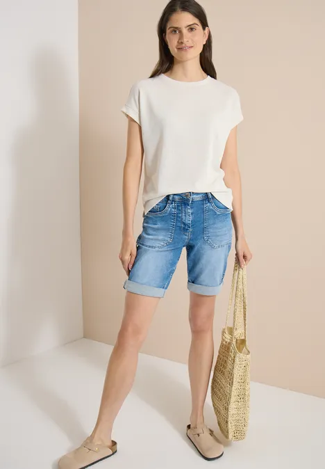Frau trägt weißes T-Shirt, Jeans-Shorts, Basttasche und Sandalen vor neutralem Hintergrund.