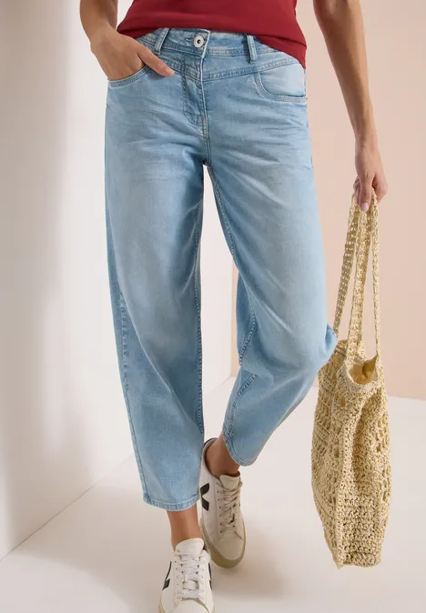Person trägt hellblaue Jeans, weiße Sneaker und hält eine gewebte Tasche.