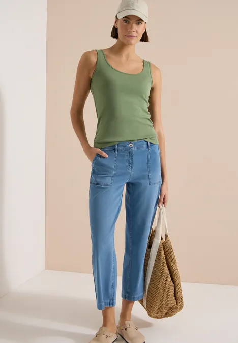 Frau mit grünem Top und blauen, locker sitzenden Jeans trägt eine geflochtene Tasche vor neutralem Hintergrund.