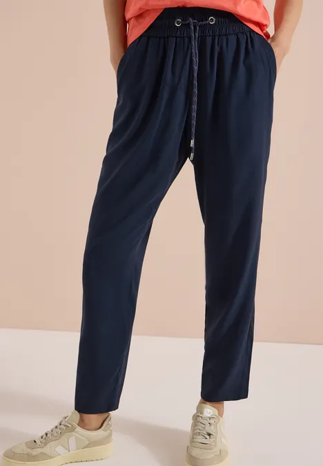 Pantalon Mid Waist Slim Leg en coupe loose universal blue Jambes portant un pantalon de jogging bleu marine à cordon et des baskets claires sur fond beige.