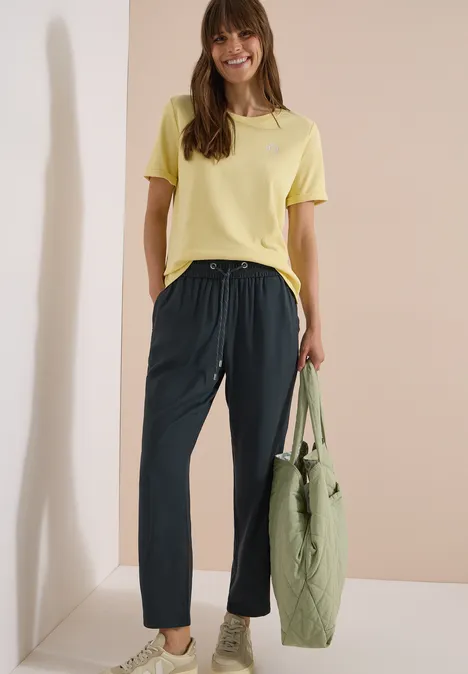 Frau trägt gelbes T-Shirt, schwarze Hose und hält eine grüne Tasche vor beigem Hintergrund.