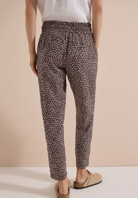 Abgeschnittenes Bild von einer Person in leopardgemusterten Stoffhosen und Slip-on-Schuhen vor beigem Hintergrund.