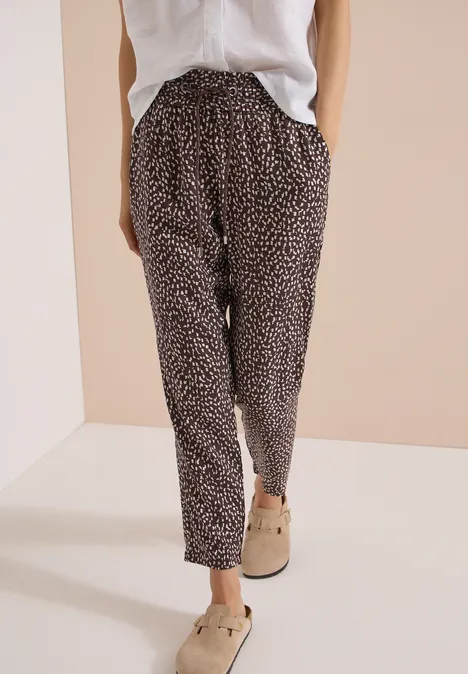 Ein Modell trägt eine locker sitzende schwarze Hose mit weißem Tupfenmuster, weiße Bluse und beige Slipper.