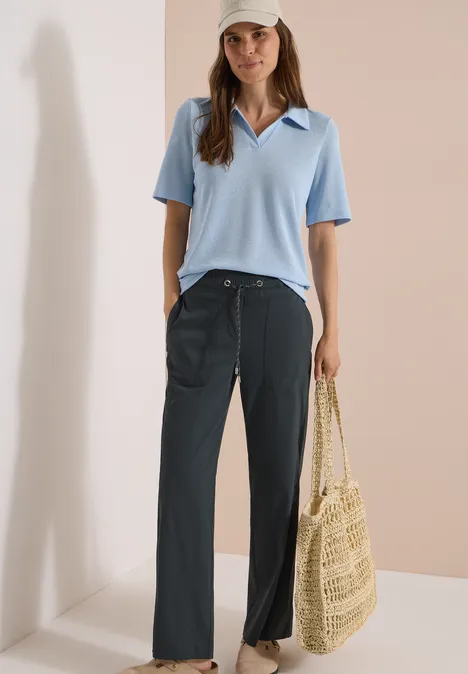 Frau trägt hellblaues Polo, weite dunkle Hose und hält eine Strohtasche.