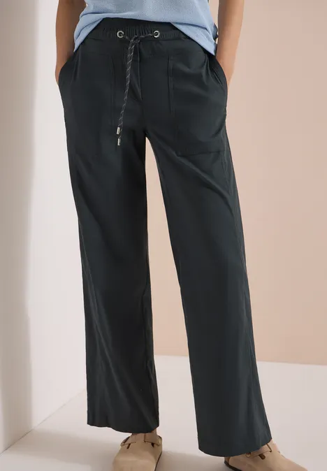 Locker geschnittene, dunkelgraue Hose mit Tunnelzug und aufgesetzten Taschen, kombiniert mit braunen Schuhen.