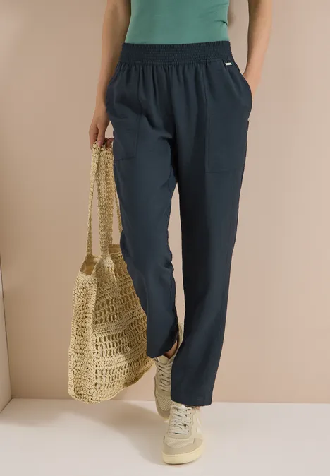 Mid Waist Slim Leg Hose im Loose Fit palm breeze green Frau trägt locker sitzende dunkelblaue Hose mit Taschen, helle Sneaker und hält einen geflochtenen Korbbeutel.