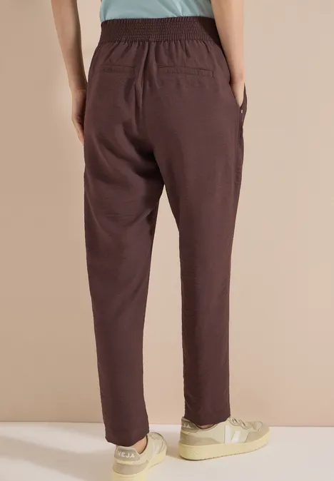 Mid Waist Slim Leg Hose im Loose Fit brown cacao Rückenansicht einer Person in locker sitzenden braunen Hosen und hellen Sneakers.