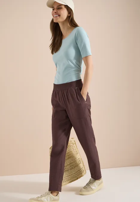 Mid Waist Slim Leg Hose im Loose Fit brown cacao Frau in hellblauem T-Shirt, dunkelroter Freizeithose und Strohtasche, entspannt stehend.