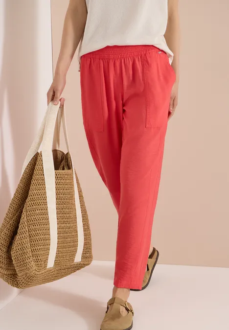 Mid Waist Slim Leg Hose im Loose Fit coral Person trägt rote Leinenhose, weiße Bluse, Basttasche und Sandalen vor hellem Hintergrund.