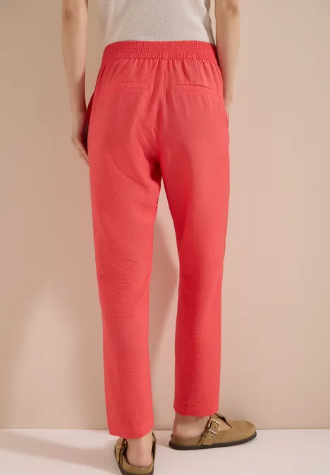 Mid Waist Slim Leg Hose im Loose Fit coral Person steht vor beiger Wand und trägt eine korallrote Hose mit grünen Pantoffeln.