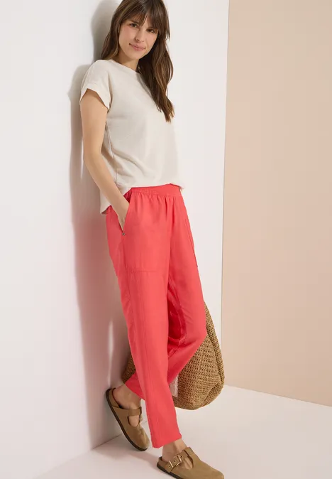 Mid Waist Slim Leg Hose im Loose Fit coral Frau lehnt an Wand, trägt beiges Top, korallrote Hose und Sandalen, hat eine geflochtene Strohtasche.