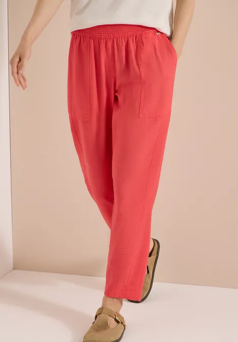 Mid Waist Slim Leg Hose im Loose Fit coral Person trägt lässige, korallrote Leinenhose mit Gummibund und seitlichen Taschen, kombiniert mit beigen Sandalen.