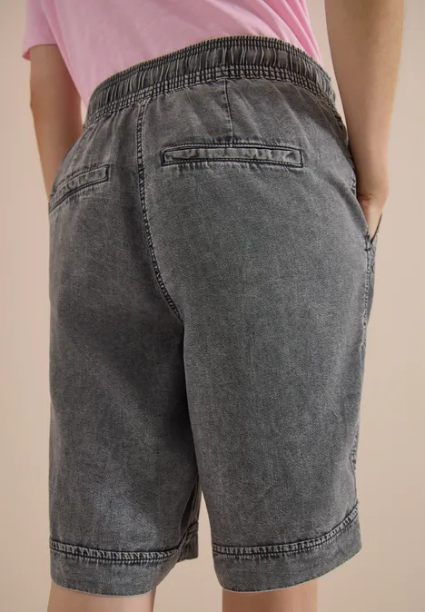 Een persoon draagt grijze denim bermudashort met elastische tailleband, achter- en zijzakken, staat met handen in de zakken.