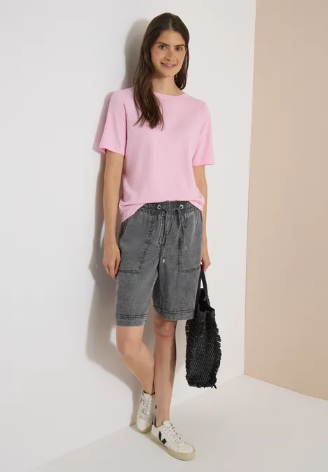 Een vrouw in een roze T-shirt, grijze bermudashorts, witte sneakers en een zwarte tas.