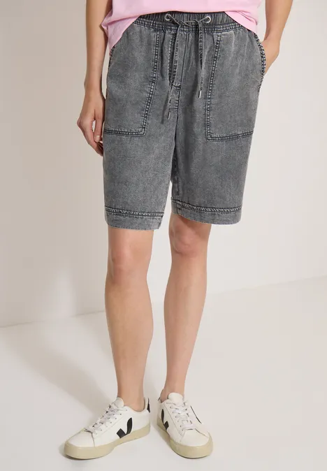 Persoon in grijze spijkershorts met zakken en witte sneakers tegen een neutrale achtergrond.