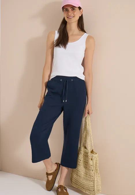Pantalon 3/4 à jambes larges en mousseline universal blue Une femme porte un top blanc, des culottes bleu marine, des sandales et tient un sac en paille.