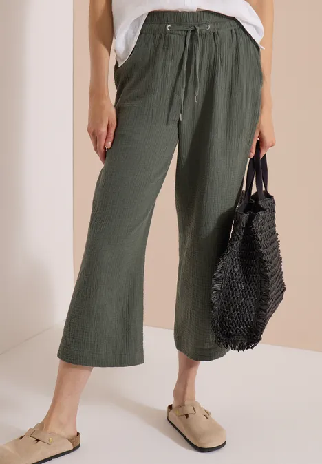 Frau trägt olivgrüne weite Culotte-Hose mit Tunnelzug, beigen Mules und schwarze Netz-Tasche