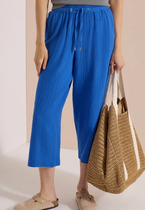 Eine Person trägt knöchellange, weite, blaue Hose mit Kordelzug, flache Sandalen und hält eine Strohtasche.