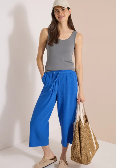 Frau in grauem Tanktop und blauer Culotte, Sandalen und geflochtener Tasche vor hellem Hintergrund.