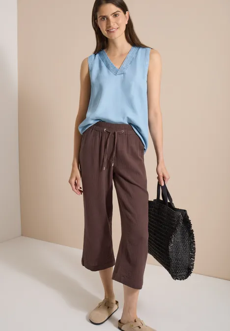Frau mit ärmellosem hellblauem V-Ausschnitttop, brauner Culotte-Hose und geflochtener schwarzer Tasche.