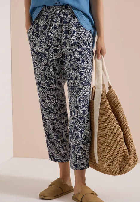 Frau trägt lässige, blaue Paisley-Hose mit Kordelzug, beigen Leder-Slides und Basttasche.