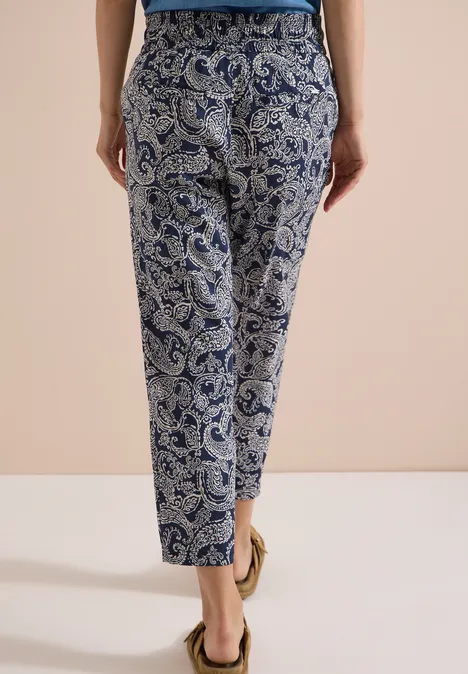 Frau von hinten in blau-weißen, gemusterten Hosen und Sandalen vor neutralem Hintergrund.