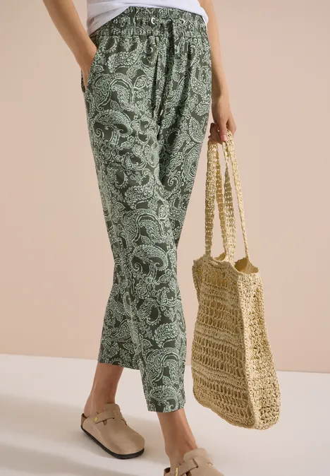 Eine Person trägt grüne, locker sitzende Paisley-Hose, beige Slipper und hält eine geflochtene Strohtasche.