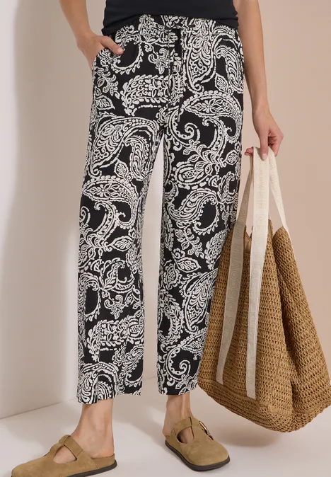Person trägt schwarz-weiß gemusterte Freizeit-Hose, bequeme Sandalen und hält eine große Strohtasche.