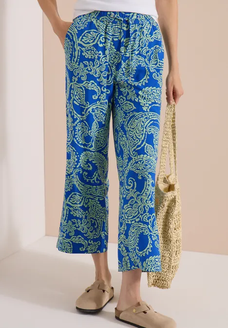 Blaue Hose mit weitem Bein und grünen Paisley-Muster, kombiniert mit Basttasche und goldenen Schuhen.