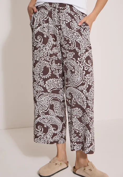 Braune, weite Hose mit weißen Paisley-Prints und Taschen, kombiniert mit beigen Pantoletten.